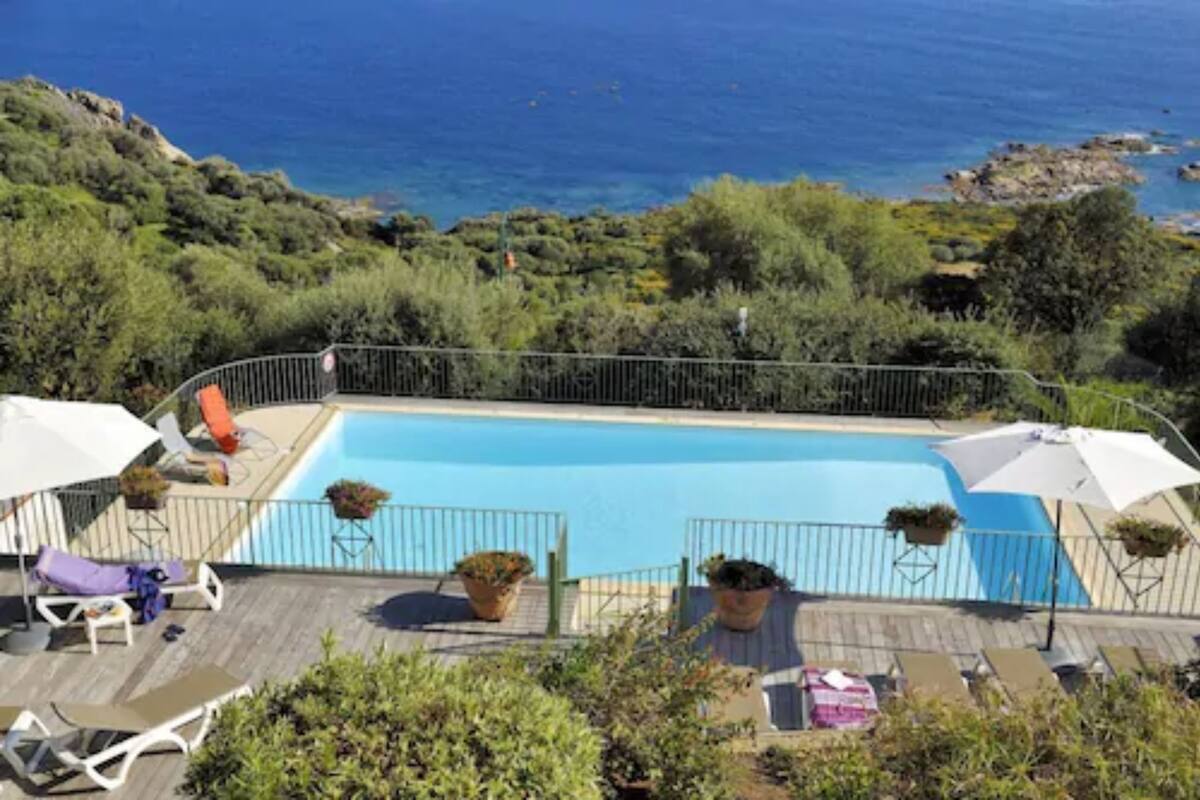 Piscina con vista al mar, rodeada de vegetación y sillas de terraza.