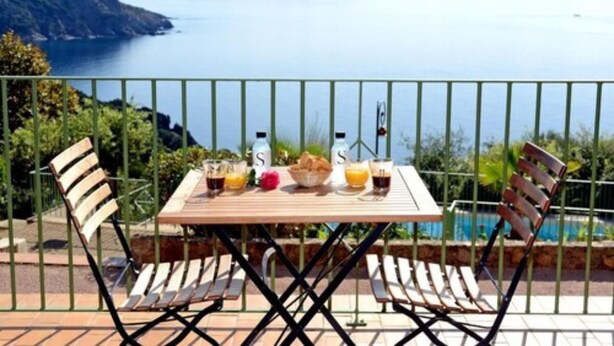 A table with two chairs, a bottle of water, a bottle of juice, and a basket of bread on a terrace with a view of a lake and mountains. - Résidence Roc e Mare (Cargese)