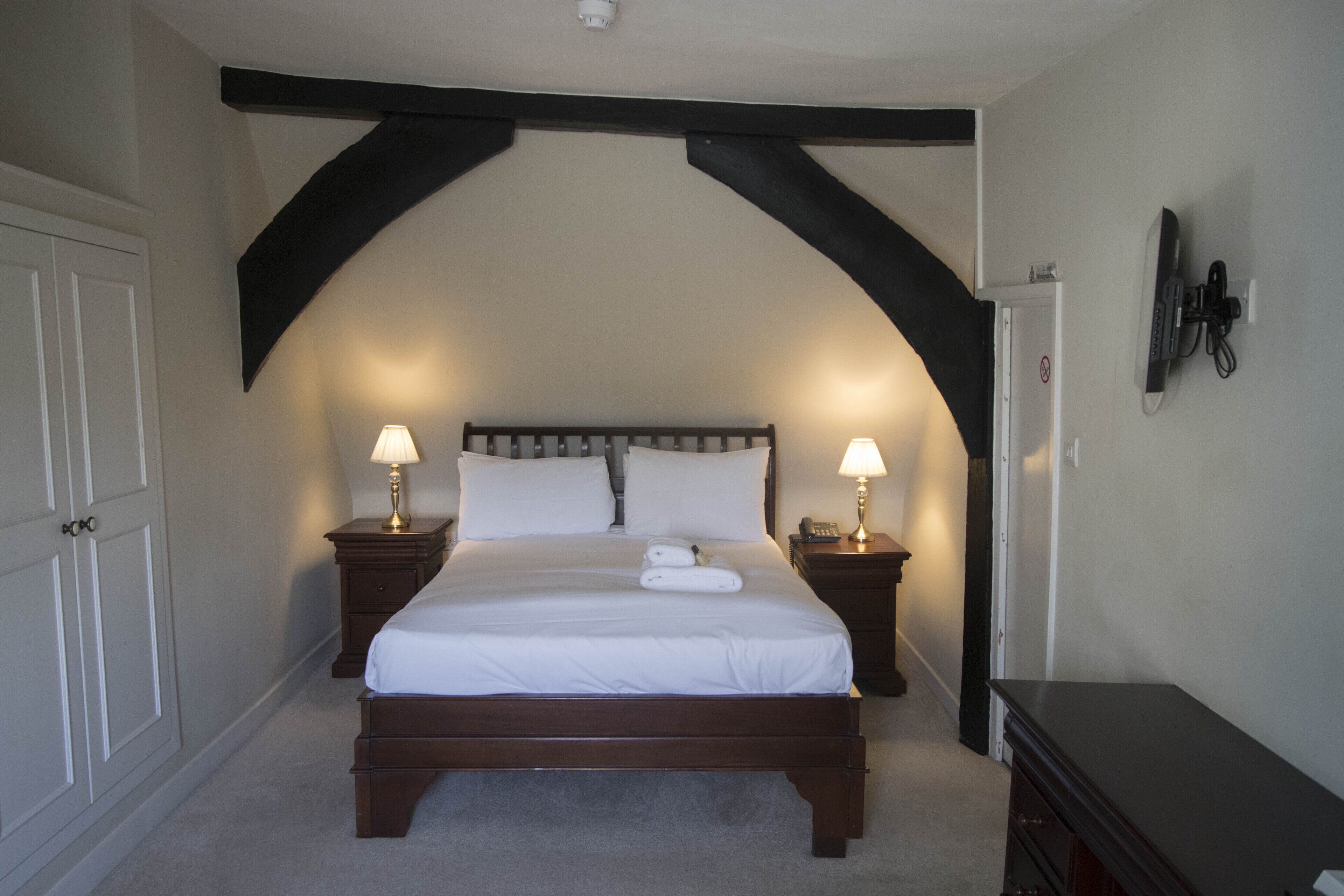 A bedroom with a large bed, two bedside tables with lamps, a wardrobe, and a flat-screen TV mounted on the wall.