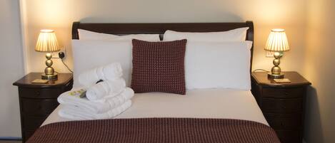Cama bem arrumada com roupa de cama branca e castanha, duas luminárias de cabeceira e cabeceira castanha.