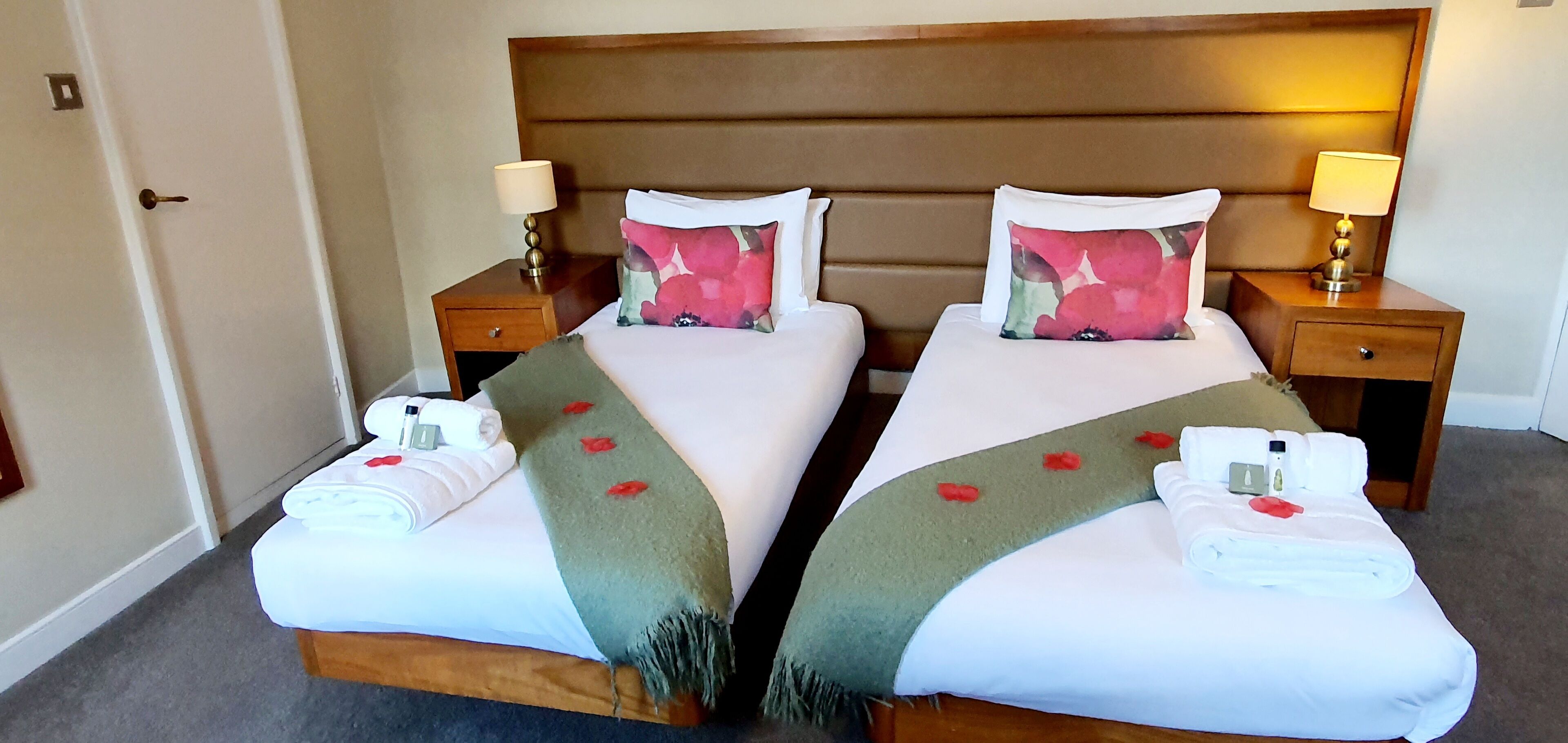 Habitación de hotel con dos camas, cada una con cabecero de madera, mesitas de noche con lámparas y toallas dobladas con cuidado sobre las camas.