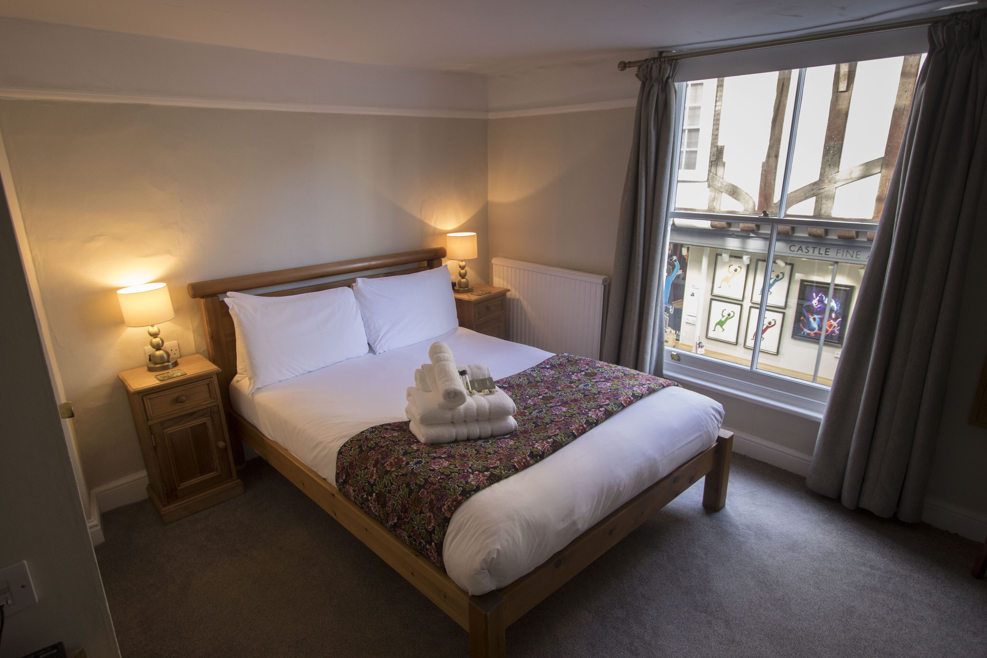 A neatly made bed with white linens, a floral-patterned bedspread, and a stack of towels on the footboard. There are two bedside lamps on wooden nightstands. A window with a view of a building with a sign reading "CASTLE FINE" is visible.
