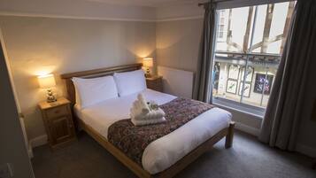 A neatly made bed with white linens, a floral-patterned bedspread, and a stack of towels on the footboard. There are two bedside lamps on wooden nightstands. A window with a view of a building with a sign reading "CASTLE FINE" is visible.