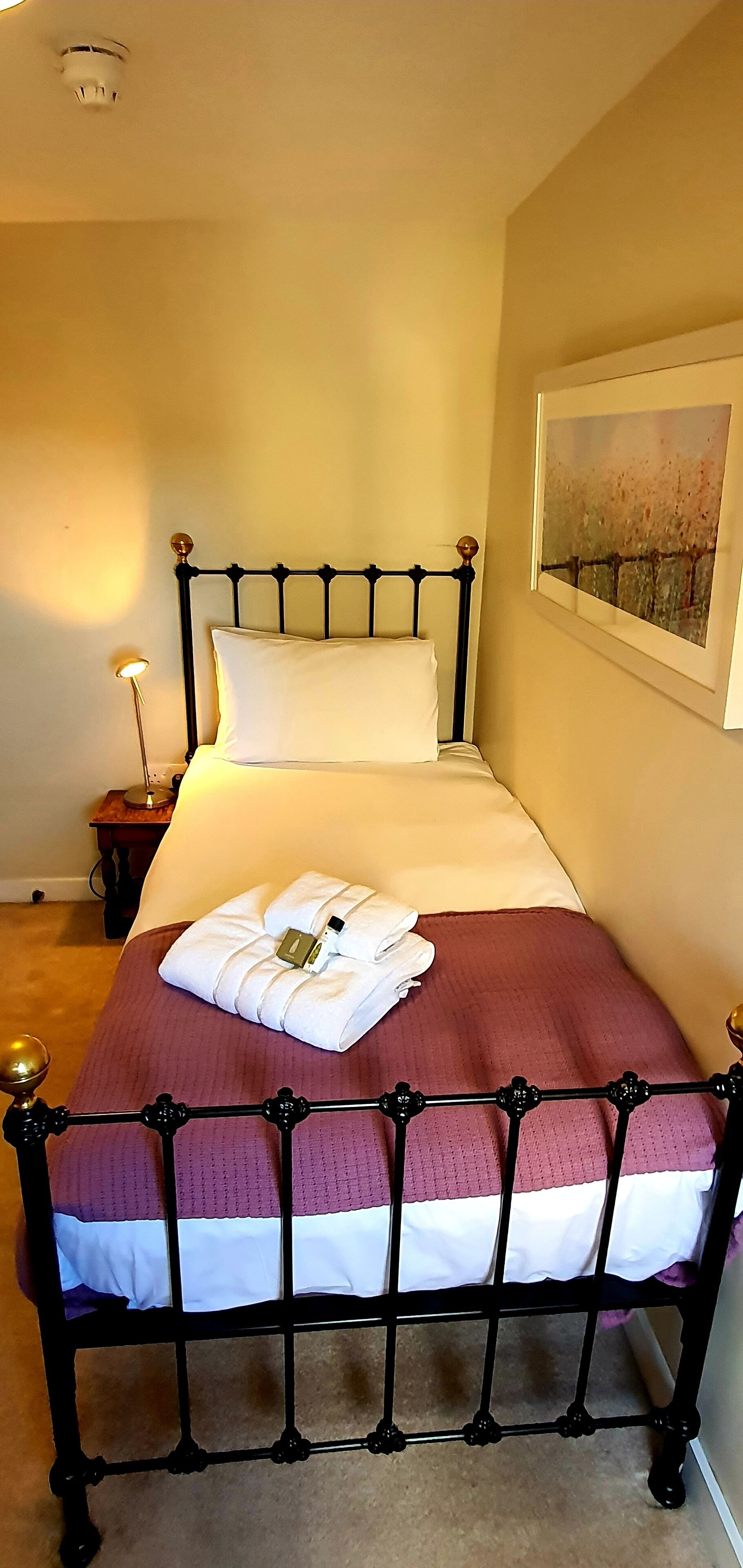 A neatly made bed with a white pillow and a folded towel, a framed artwork on the wall, and a bedside lamp.