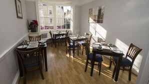 Un espace repas avec des tables et des chaises en bois, une fenêtre donnant sur des immeubles et un tableau encadré au mur.