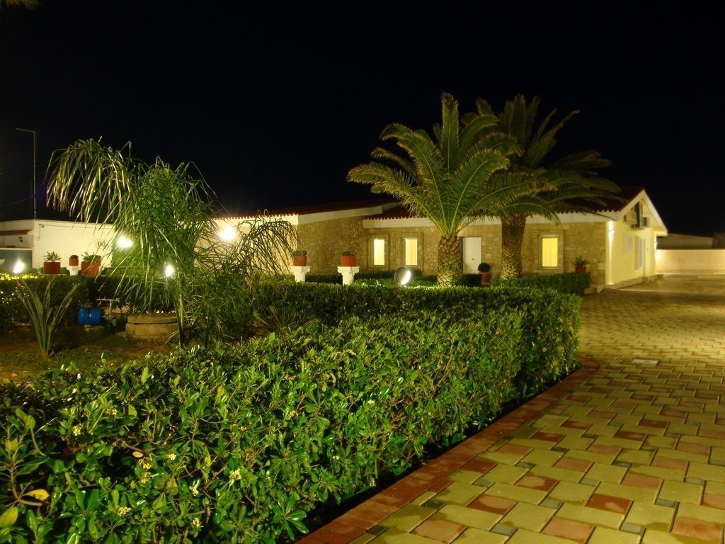 A well-lit hotel exterior at night with a paved walkway, neatly trimmed hedges, and palm trees.