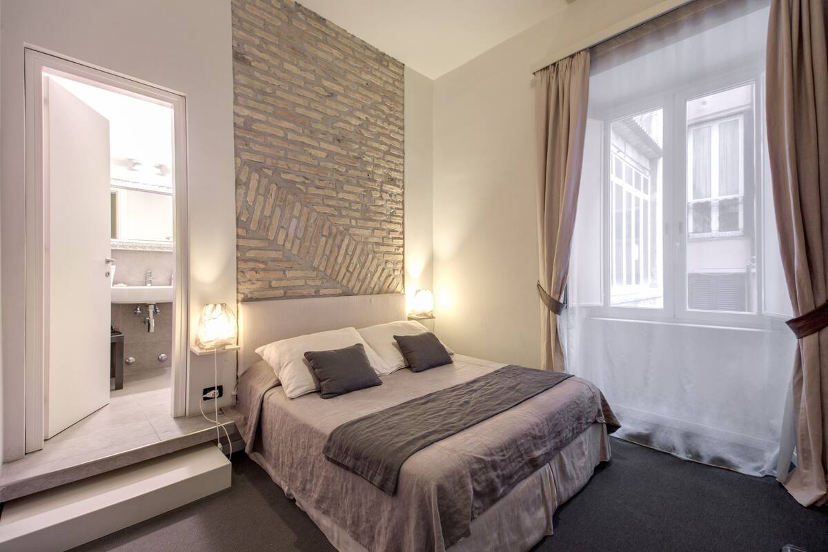 A modern bedroom with a bed, bedside lamps, a stone accent wall, a window with curtains, and a view of the outside.