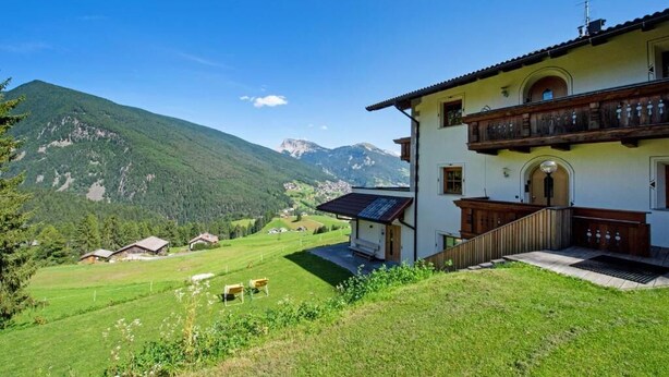 Une maison à deux étages dotée d’un balcon, entourée d’un paysage verdoyant et luxuriant, avec des montagnes en toile de fond.