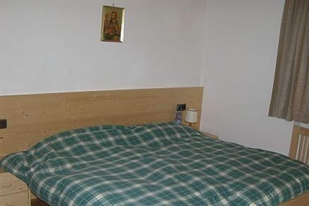 A simple room with a wooden headboard, a green plaid bedspread, a small bedside table with a lamp, and a window with a curtain.