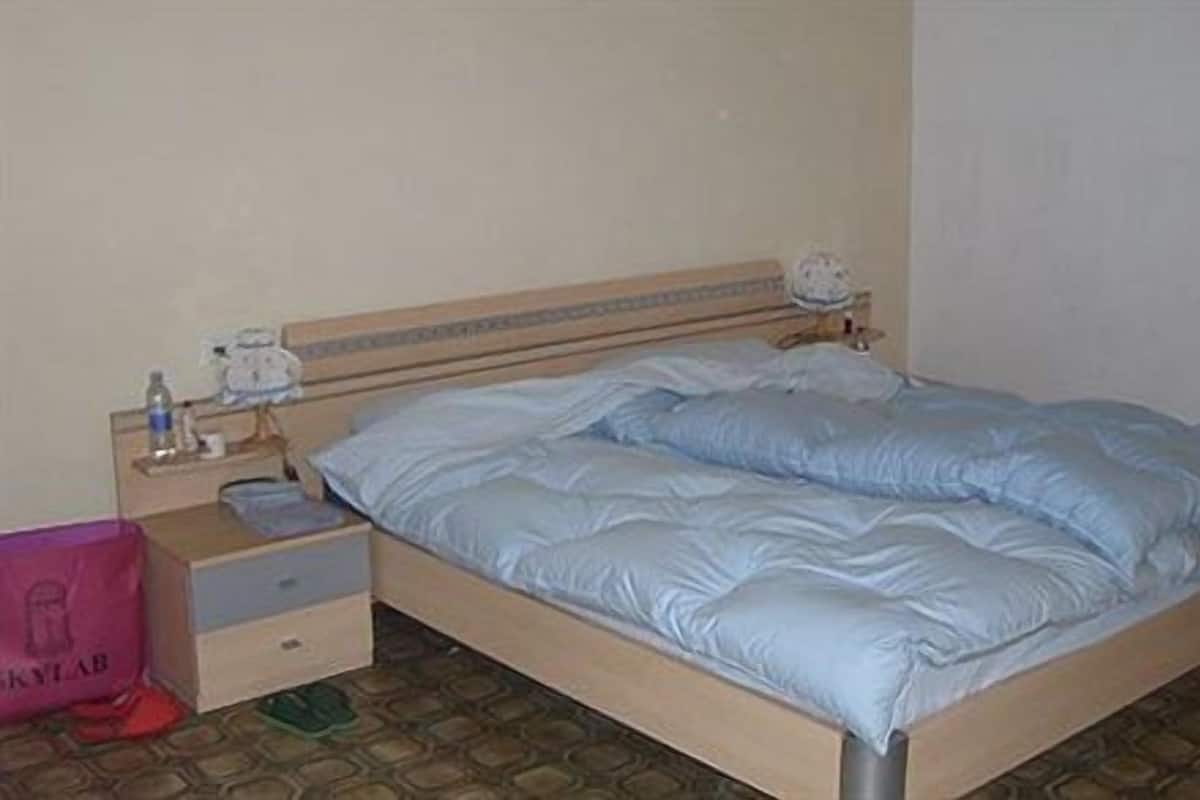 A single bed with a wooden frame and a headboard, a bedside table with a lamp, and a pink bag on the floor.
