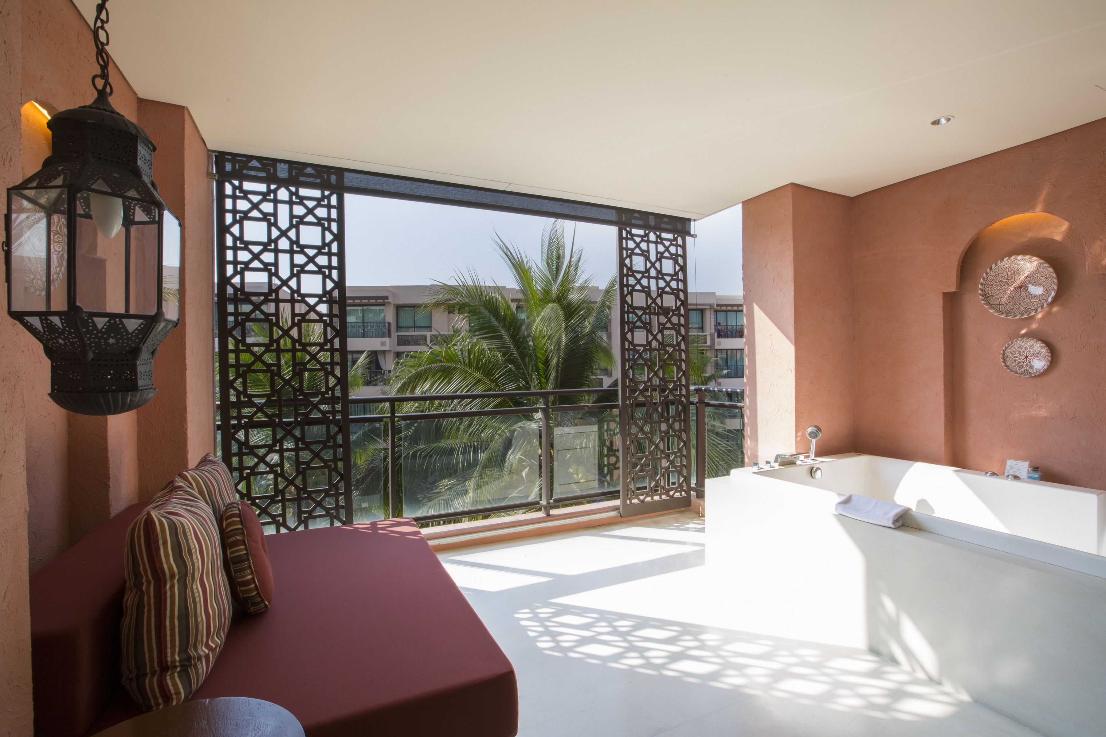 Un baño moderno con una bañera grande, una zona de estar con cojines y vistas a palmeras a través de una ventana con celosía.