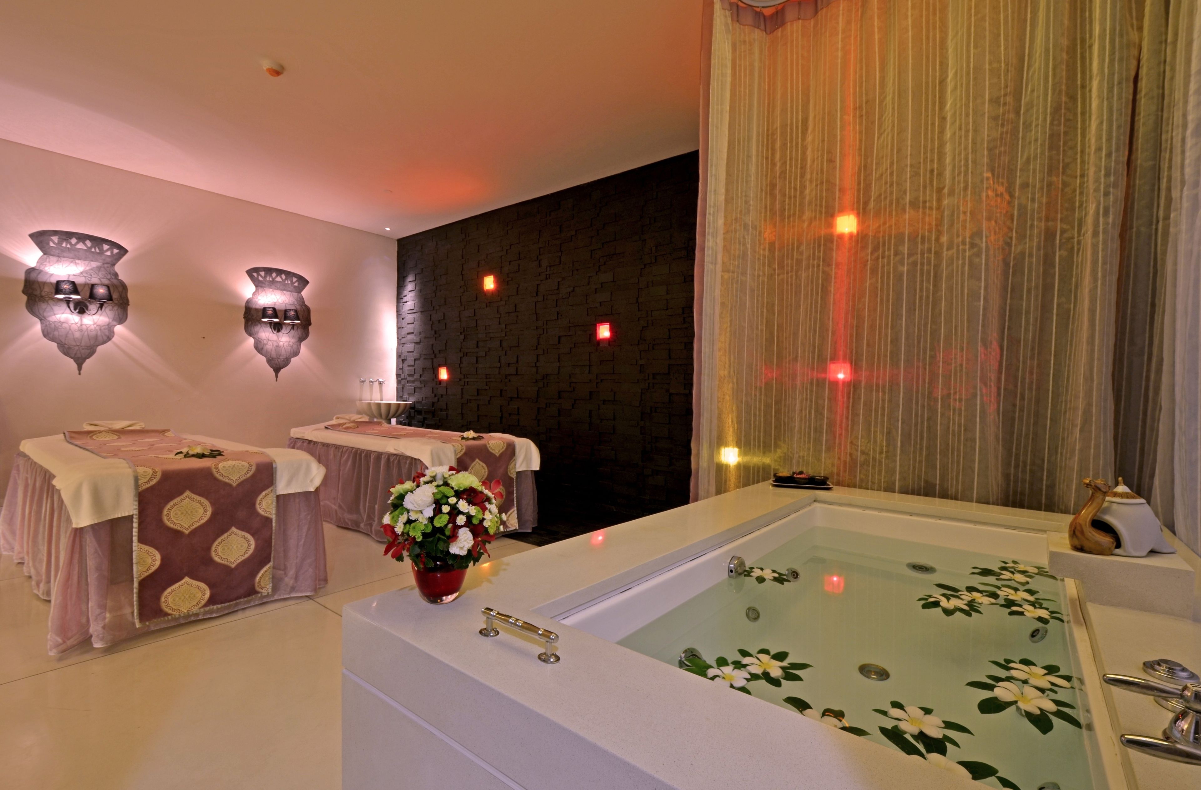 Een spa ruimte met een groot bad, twee massagetafels, wandlampen en een gordijn.