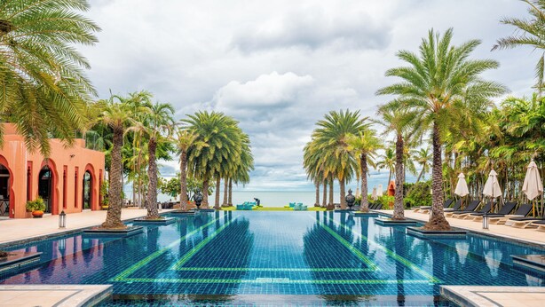 A large, clear blue infinity pool with palm trees and lounge chairs, surrounded by a red building with arches. - Marrakesh Hua Hin Resort & Spa (Hua Hin)