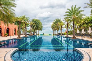 Ein großer, klarer Infinity-Pool mit Palmen und Liegestühlen, umgeben von einem roten Gebäude mit Bögen.