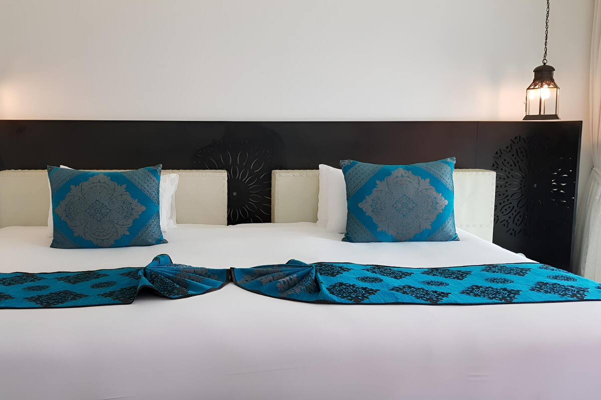 Un lit double avec une literie blanche, deux coussins décoratifs et un chemin de lit bleu à motifs.