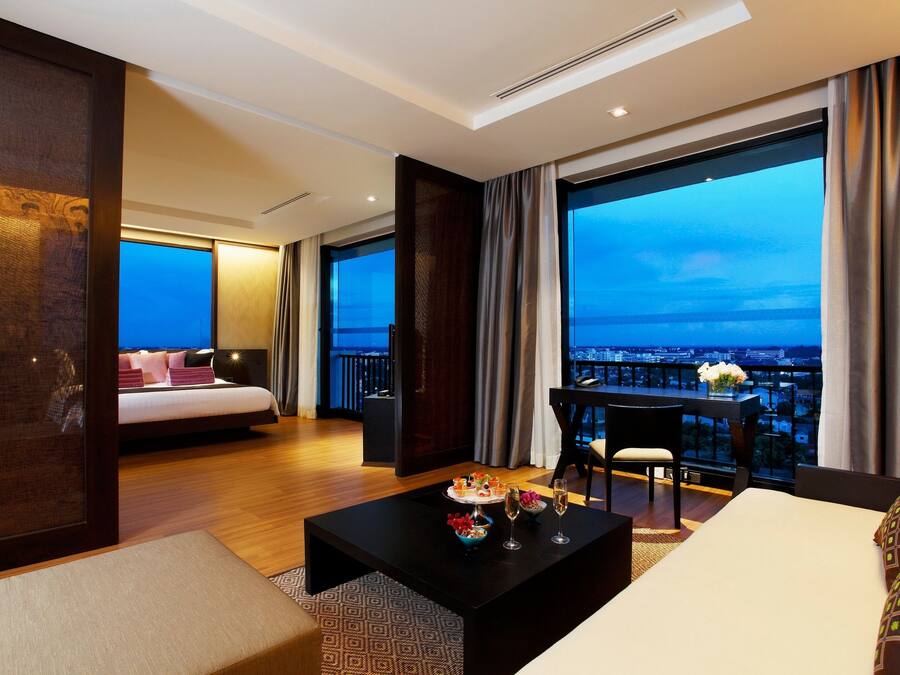 A modern hotel room with a large balcony, a bed, a dining table with a vase of flowers, and a view of the cityscape.
