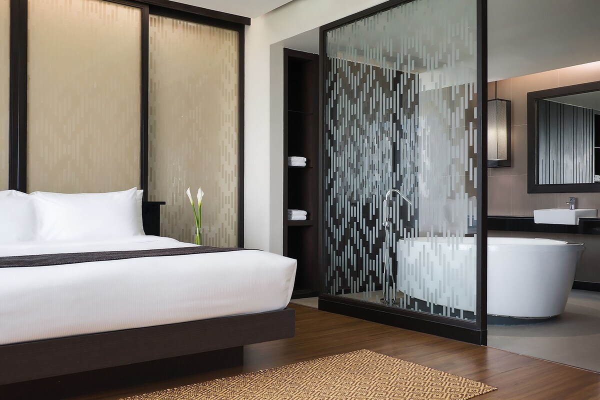 Una habitación de hotel moderna con una cama grande, un separador de vidrio, un baño con bañera y un espejo.