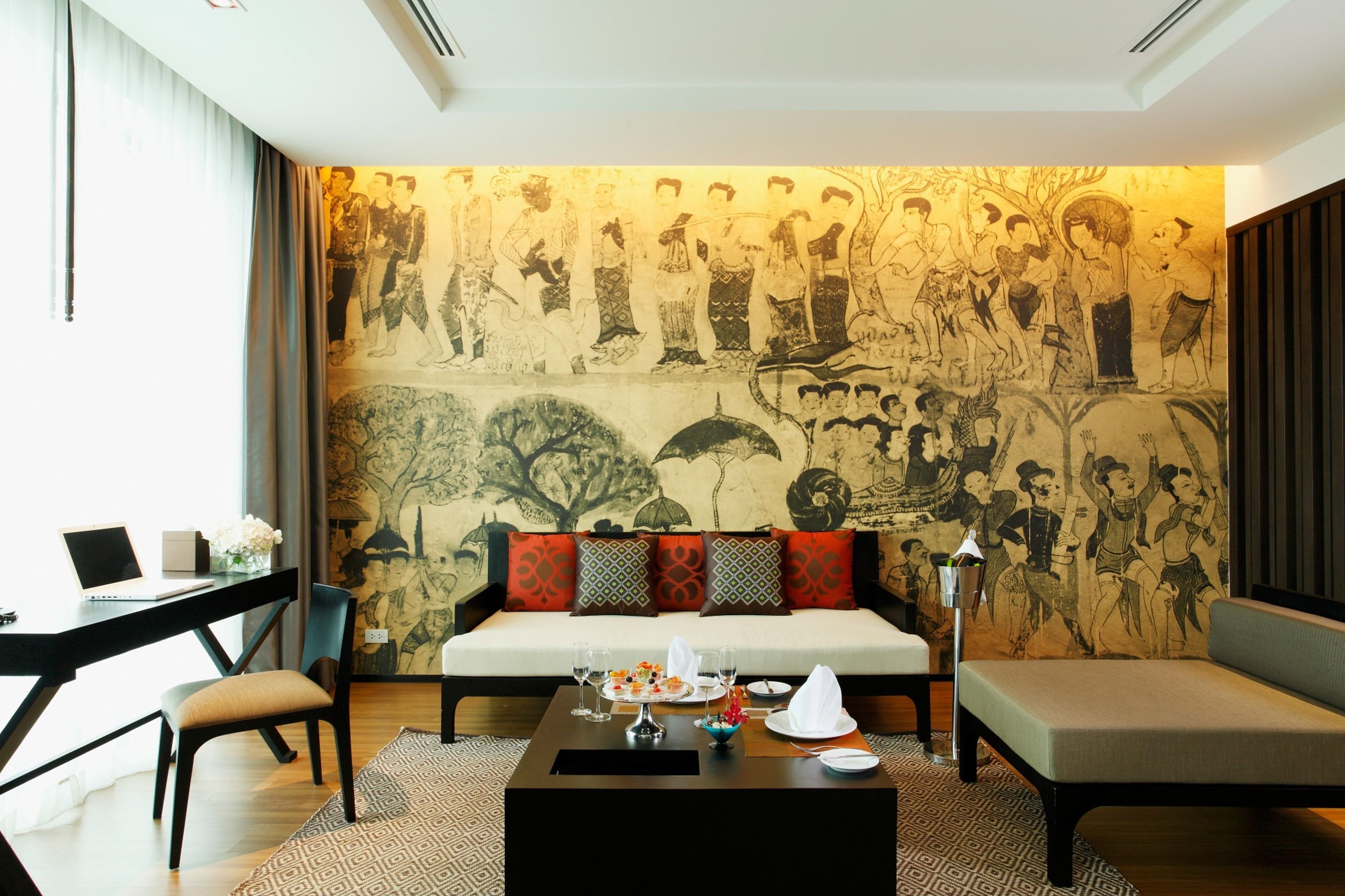 A modern living room with a traditional mural, a sofa, a coffee table, and a dining area with a laptop and flowers.