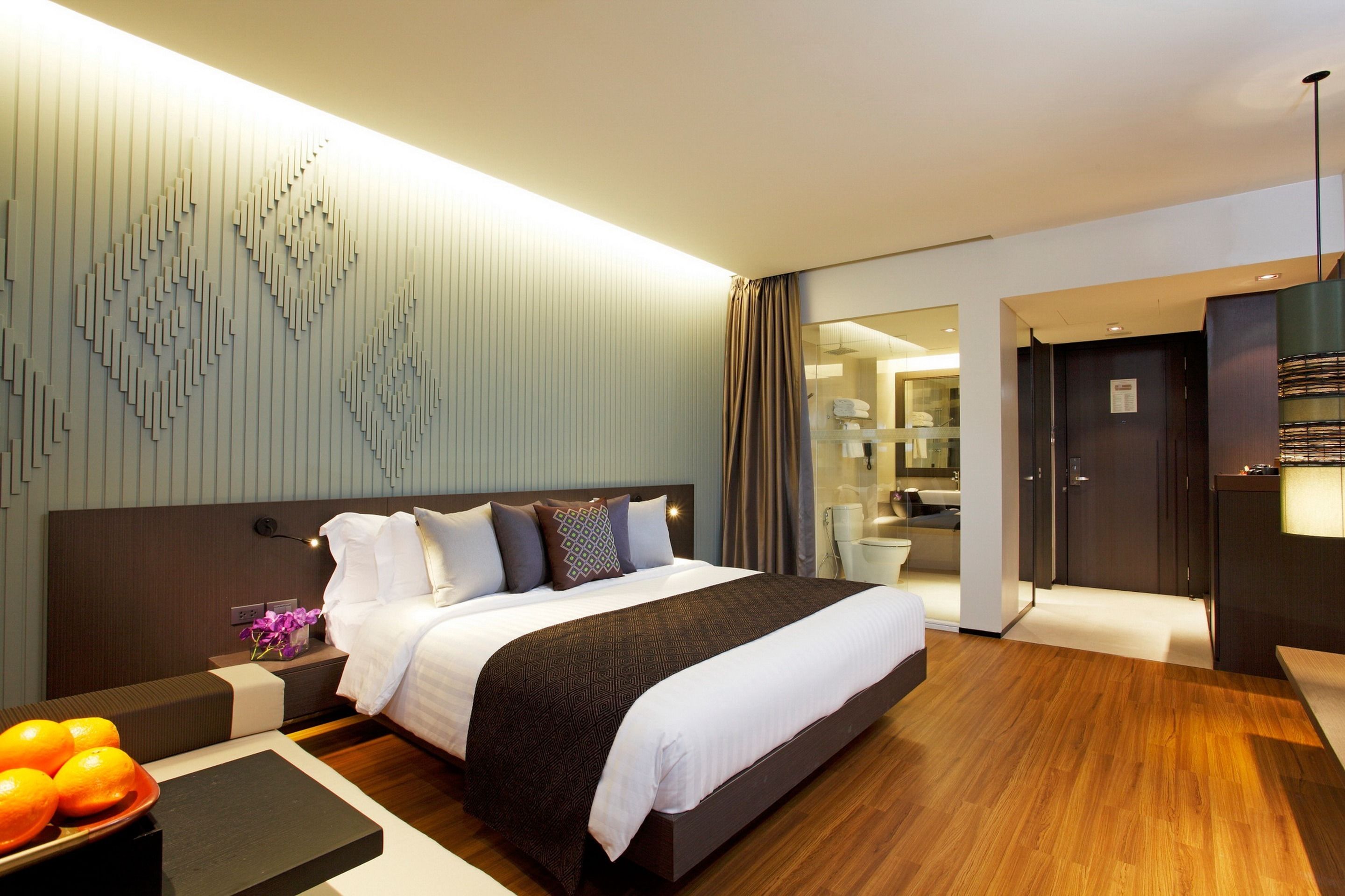 A modern hotel room with a large bed, a bedside table with a bowl of fruit, a wall-mounted light fixture, and a view into the bathroom.