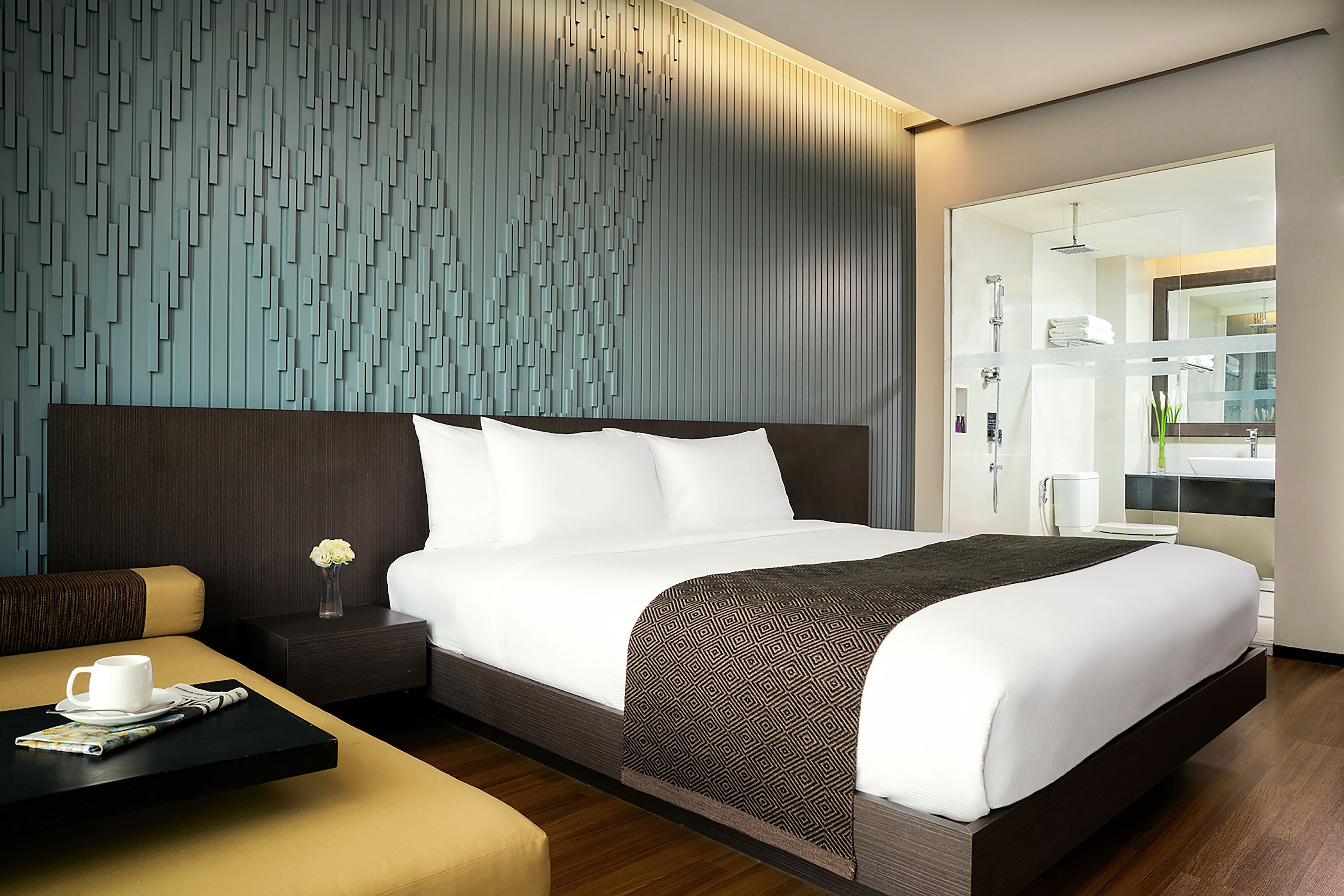 A modern hotel room with a large bed, a bedside table with a vase of flowers, a coffee table with a cup and saucer, and a view of the bathroom through a glass door.