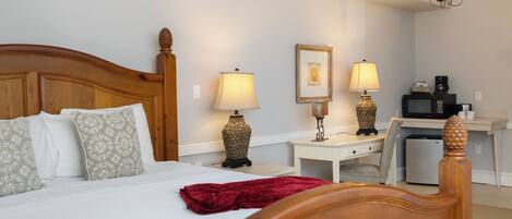 A hotel room with a wooden bed, two bedside lamps, a desk with a chair, a microwave, and a refrigerator.
