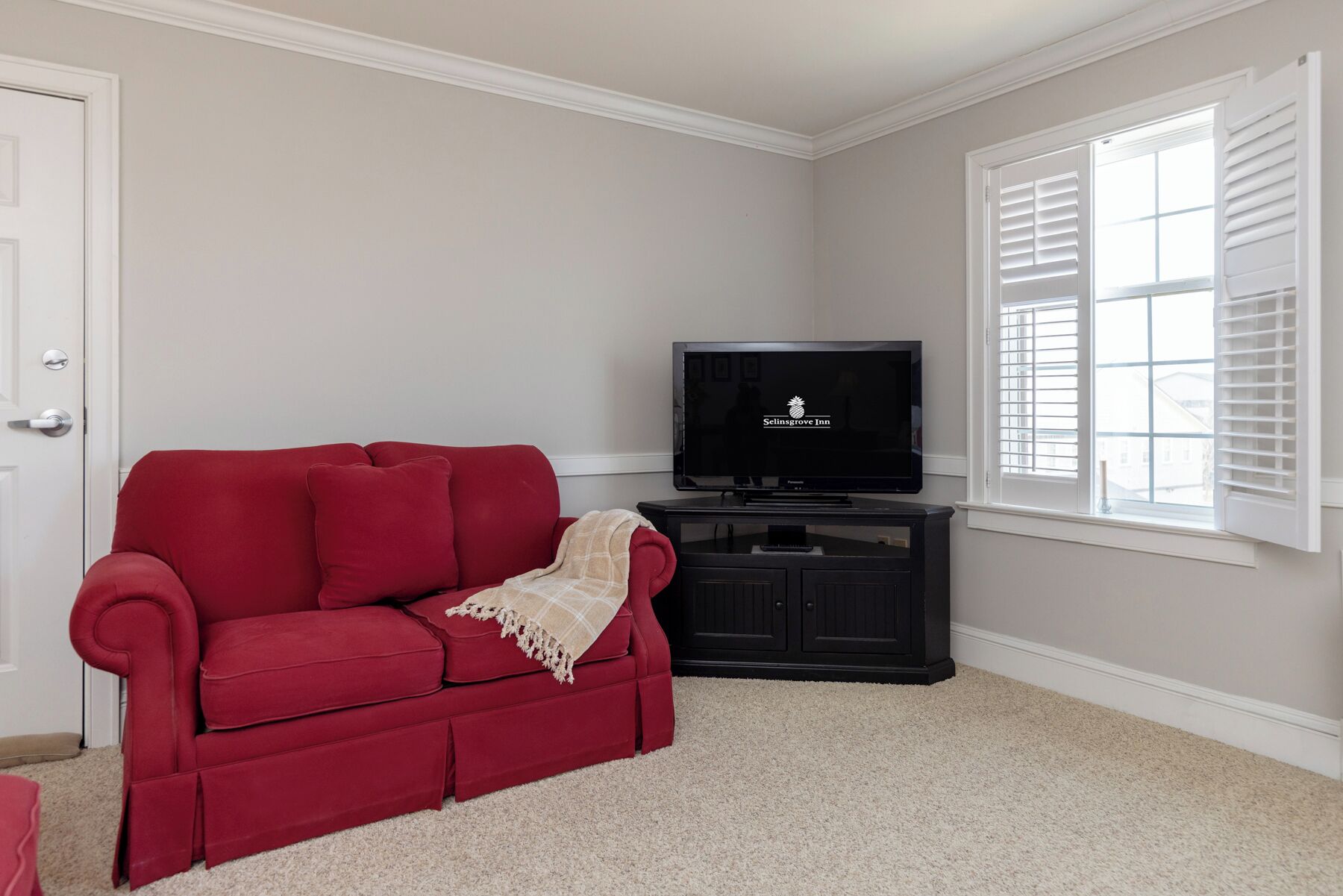 A red sofa, a television, and a window with blinds in a room with beige carpet and white walls.