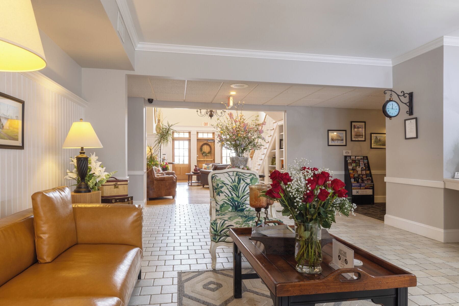 A hotel lobby with a leather sofa, a wooden coffee table with a vase of flowers, a brick-patterned floor, and a staircase in the background.