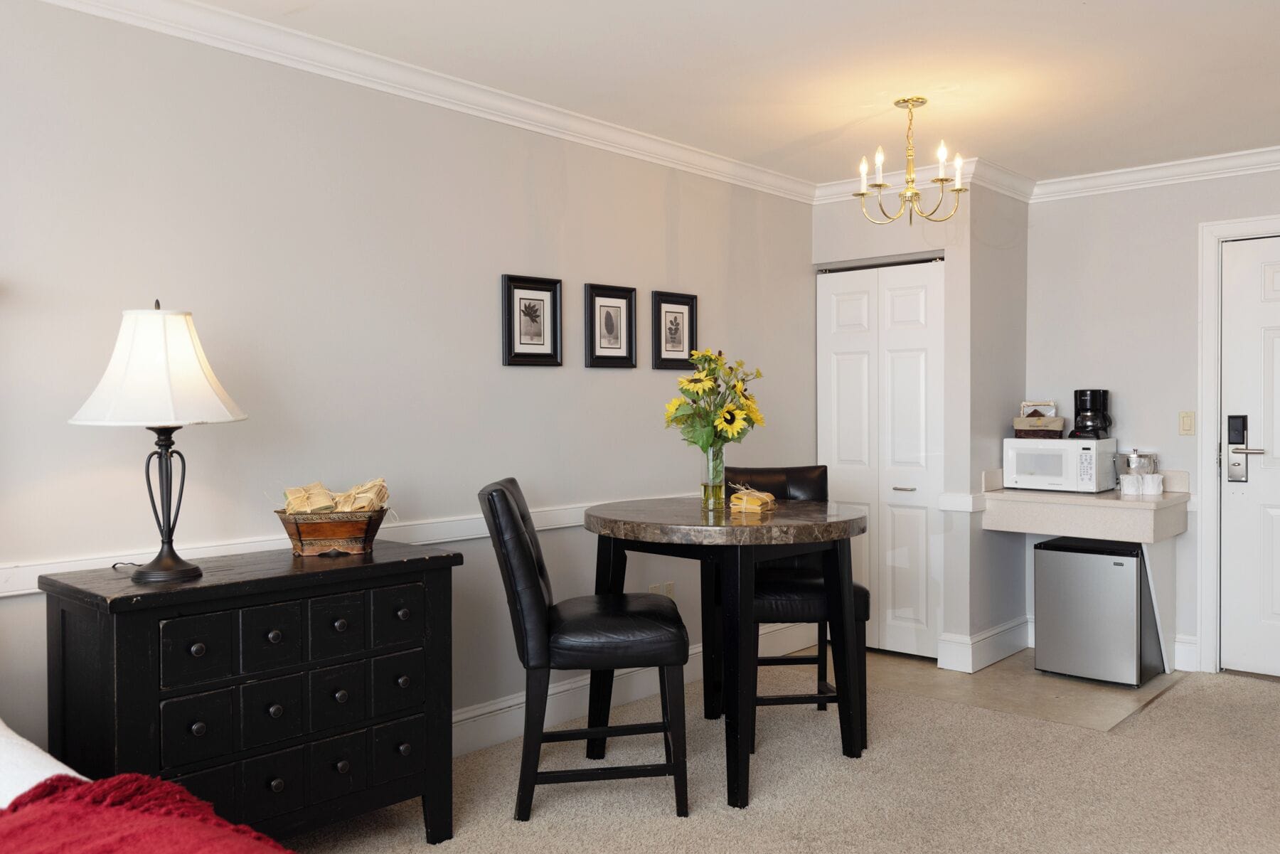 A compact hotel room with a dining table, two chairs, a lamp, a dresser, a microwave, a coffee maker, and a door.