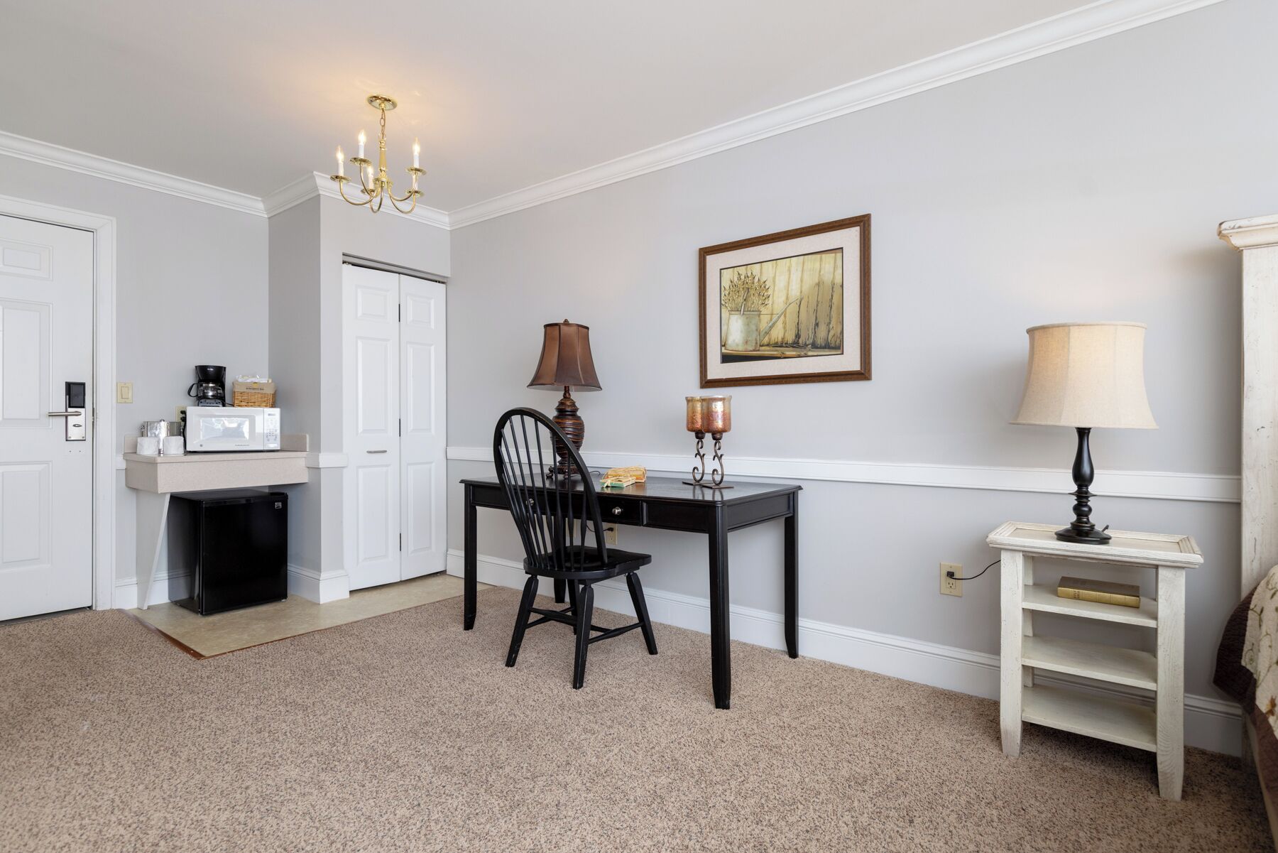 A hotel room with a dining area, a black chair, a table, a lamp, a framed picture on the wall, and a small white shelf with a book.