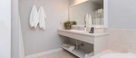 A modern bathroom with a white sink, mirror, and bathtub. There are two towels hanging on the wall and a shelf with toiletries.