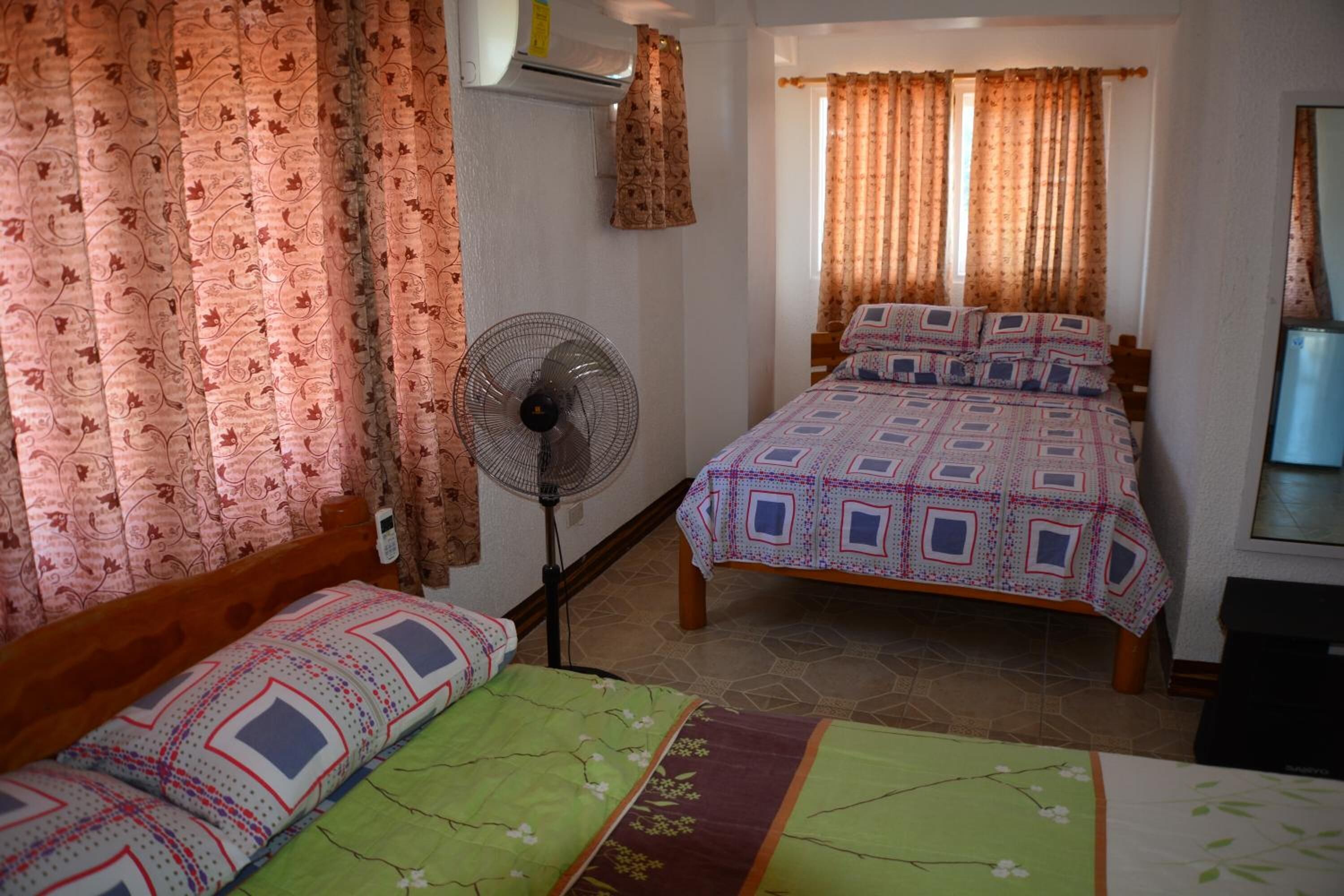 A room with two beds, a standing fan, and patterned curtains.