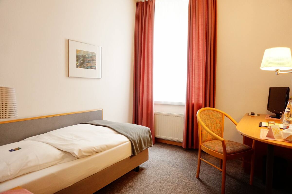 A hotel room with a bed, a chair, a desk with a computer, a lamp, and a window with curtains.