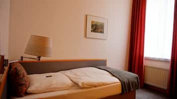 A single bed with a headboard, a bedside table with a lamp, a framed picture on the wall, and a window with red curtains.