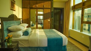 A neatly made bed with a blue and white bedspread, a wooden wardrobe, and a large window with curtains. - AG Hotel (Abu Dhabi)