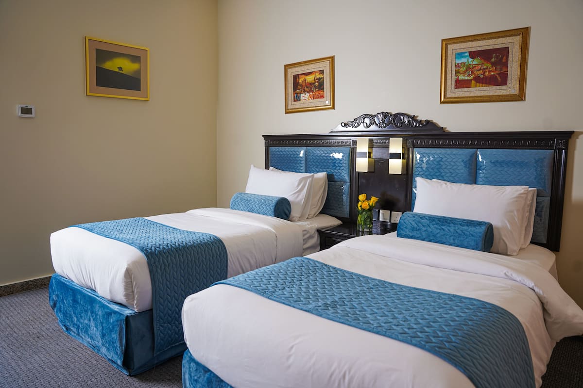A hotel room with two beds, each with blue and white bedding, a nightstand with a vase of flowers, and two framed pictures on the wall.