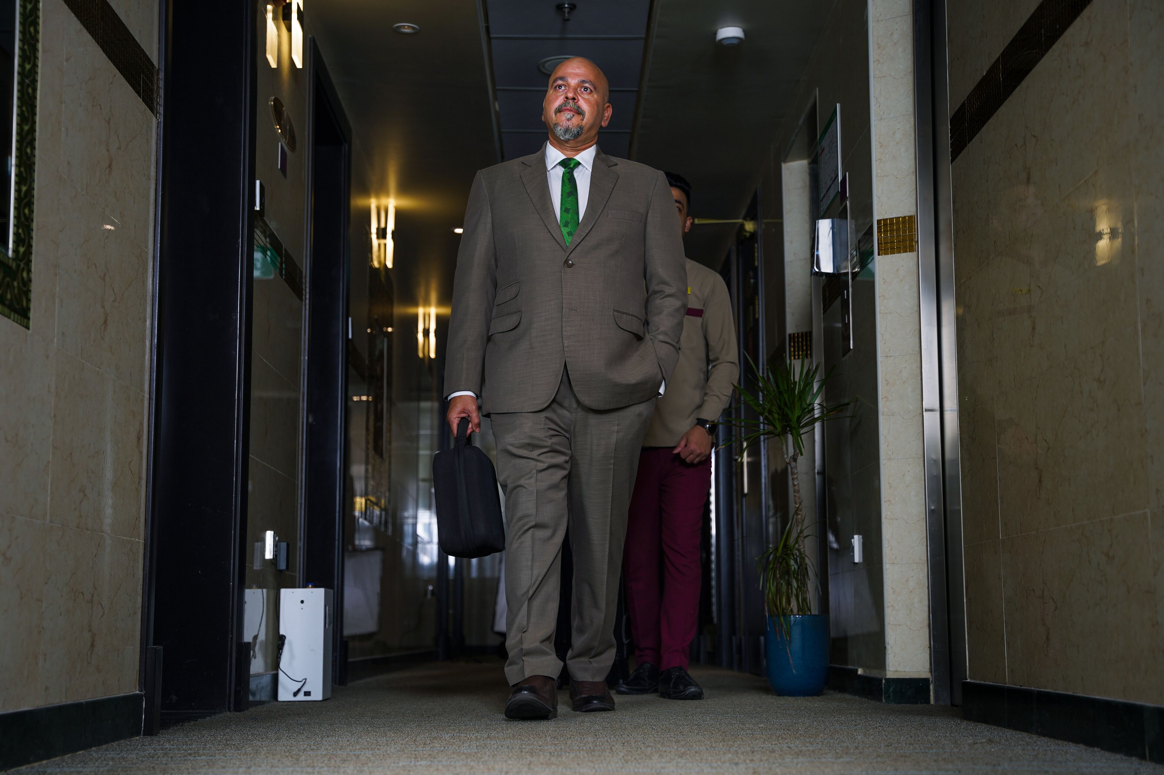 A man in a suit walking down a hallway with a briefcase, another person in the background, and a potted plant on the right.