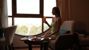 A woman sitting by a window with a plate of food and a drink, in a room with a view of greenery outside. - AG Hotel (Abu Dhabi)