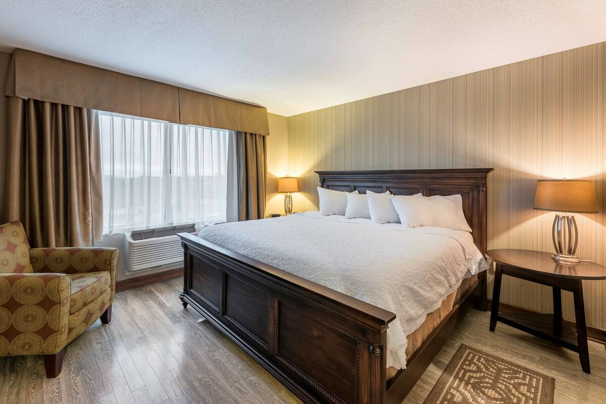 A hotel room with a large bed, a chair, a bedside table, and a lamp.