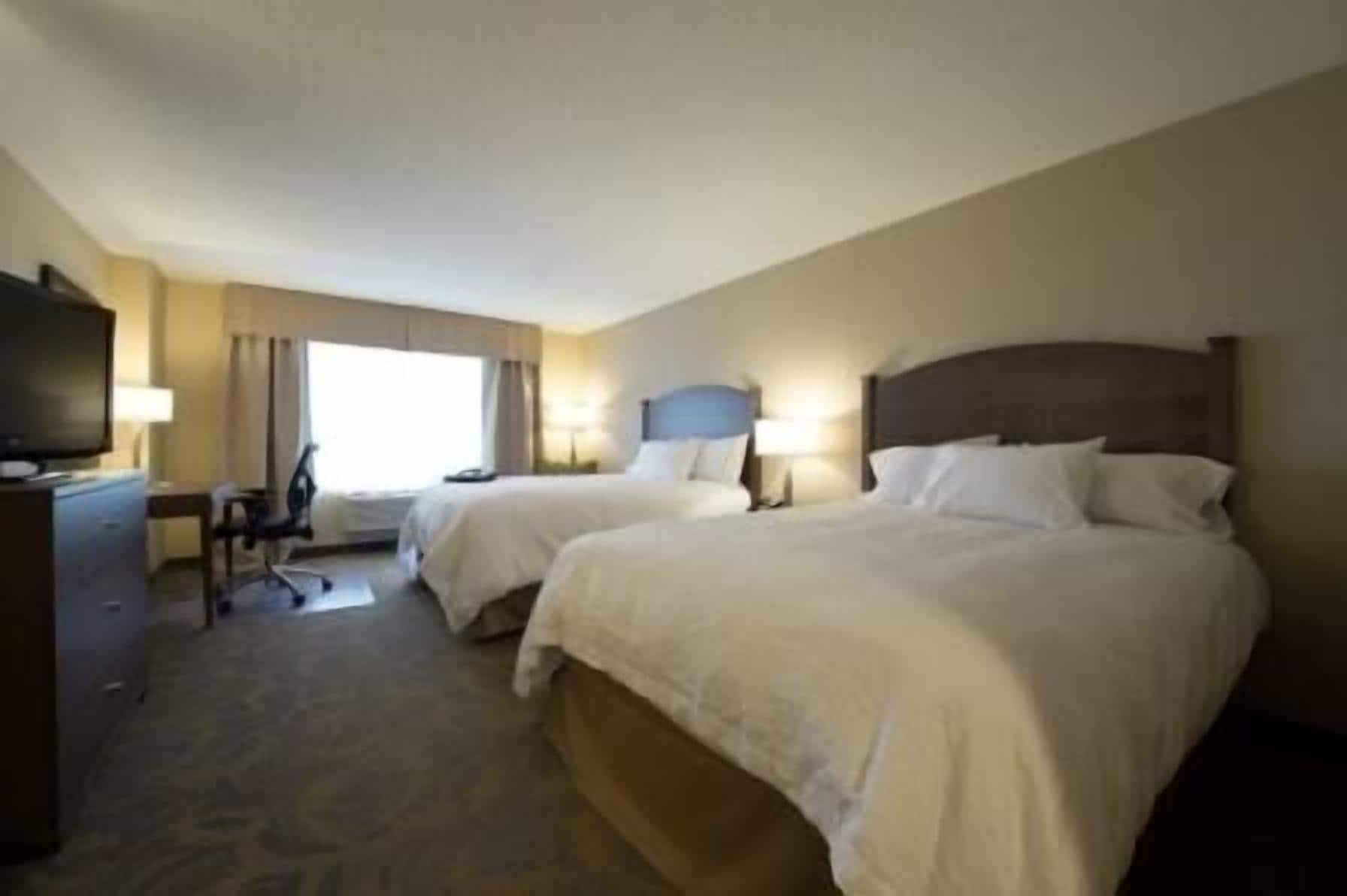 A hotel room with two beds, a desk, a chair, a television, and a window with curtains.