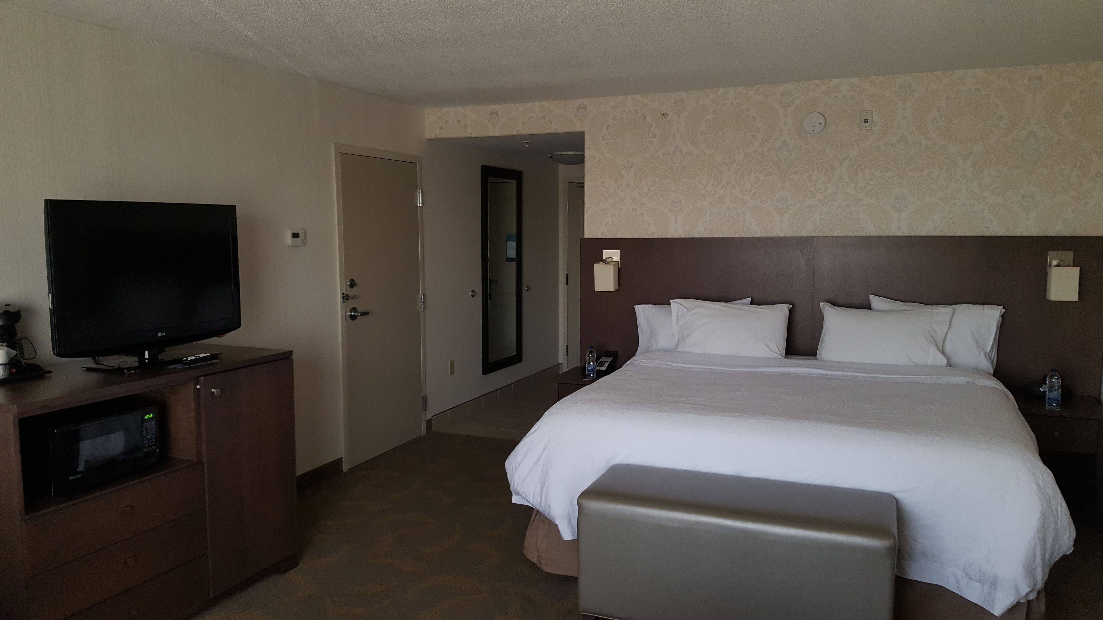 A hotel room with a large bed, a flat-screen TV, a minibar, and a bathroom door.