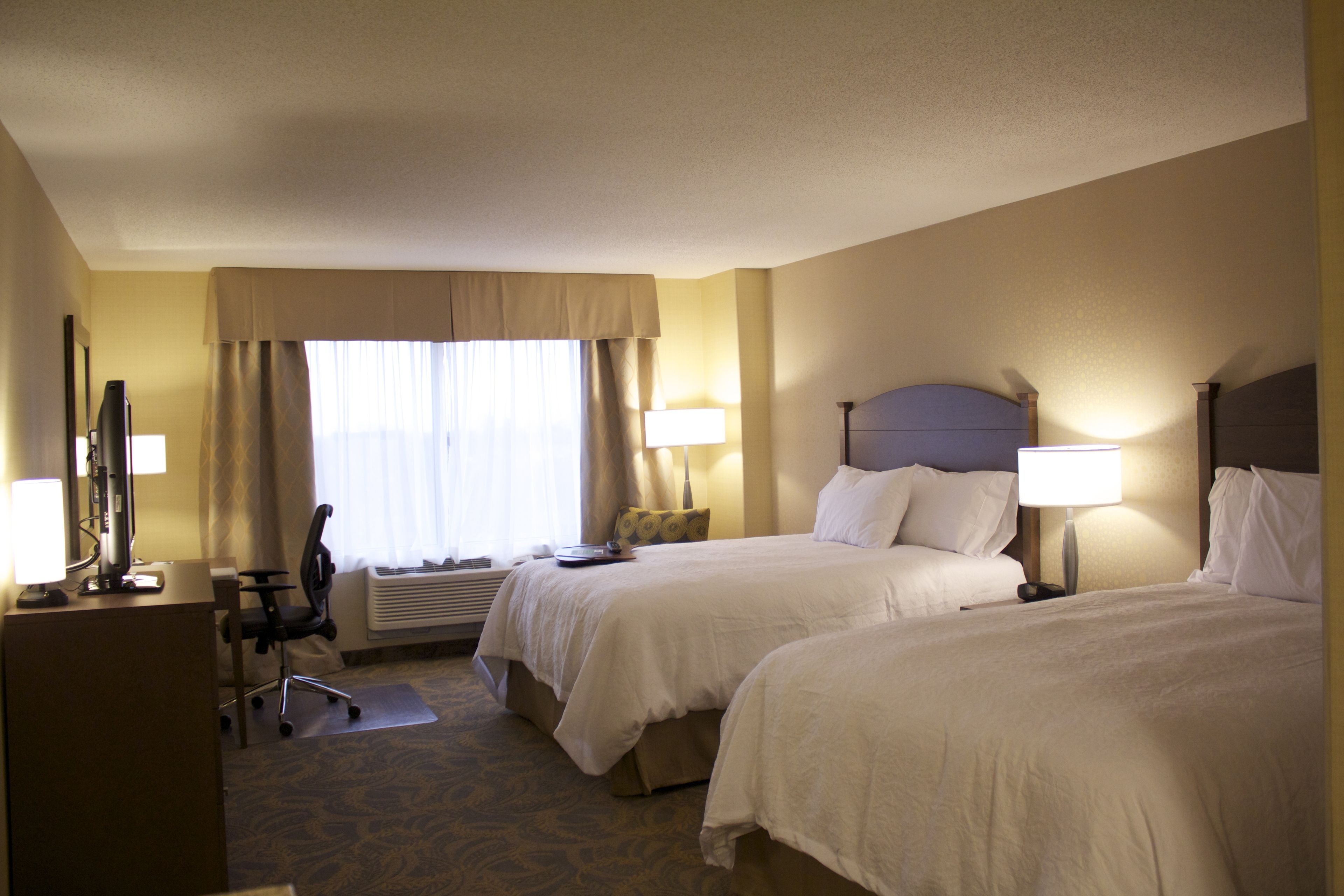 A hotel room with two beds, a desk, a chair, and a window with curtains.