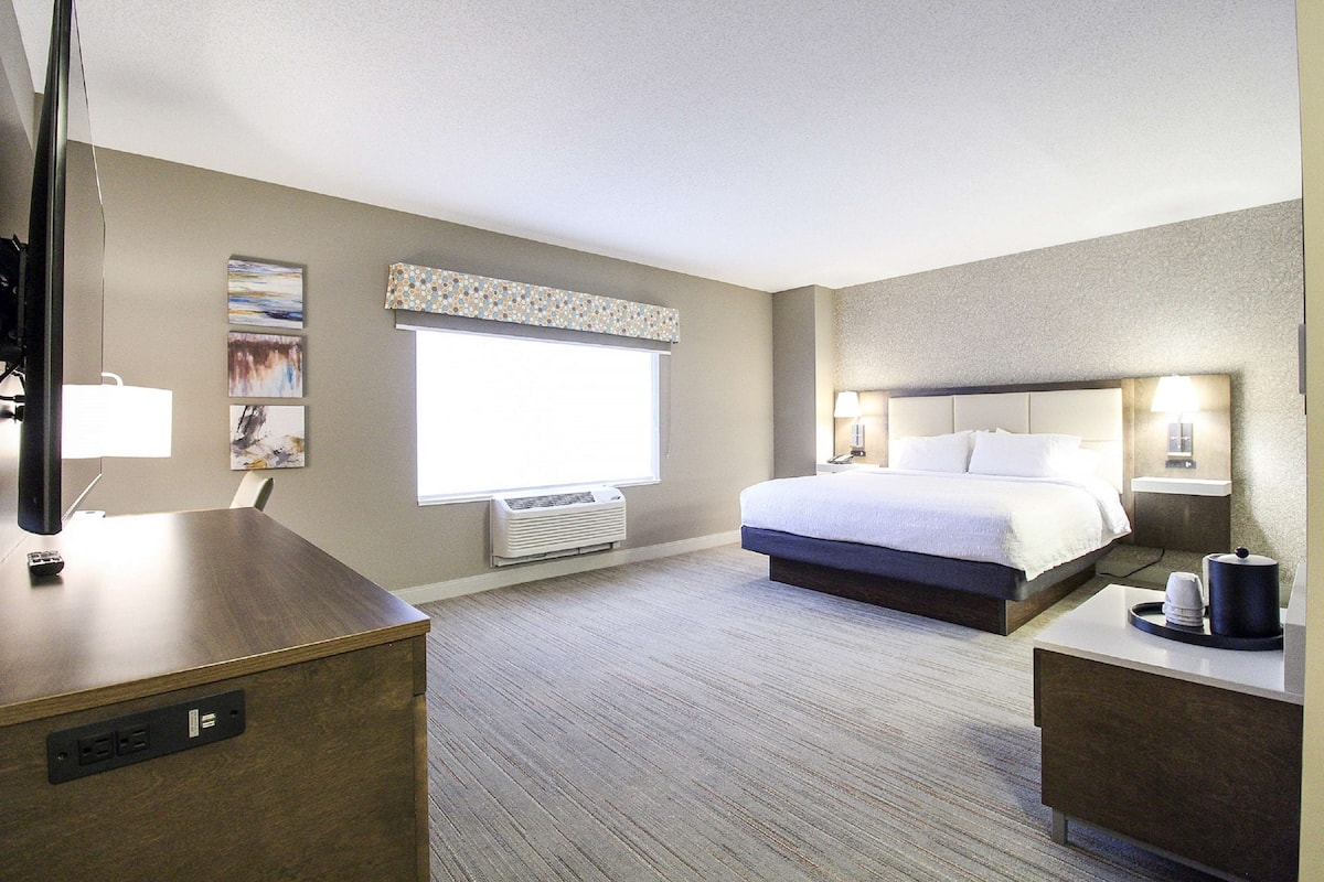 A hotel room with a bed, bedside table, lamp, air conditioning unit, and a desk with a computer.