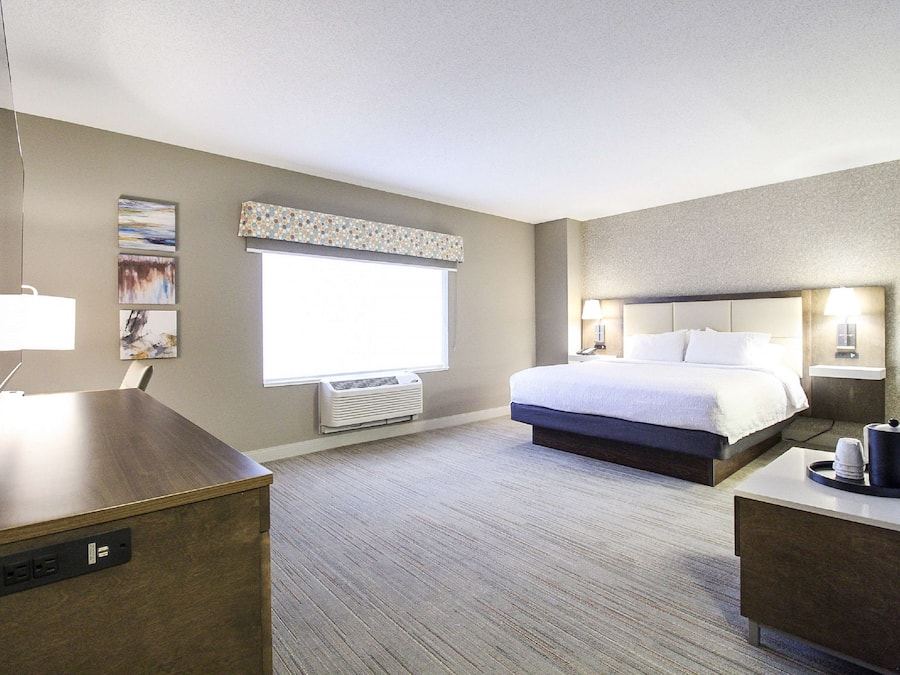 A hotel room with a bed, bedside table, lamp, air conditioning unit, and a desk with a computer.