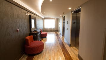 Un pasillo de hotel con piso de madera, sillas tapizadas en rojo, un espejo y un ascensor.