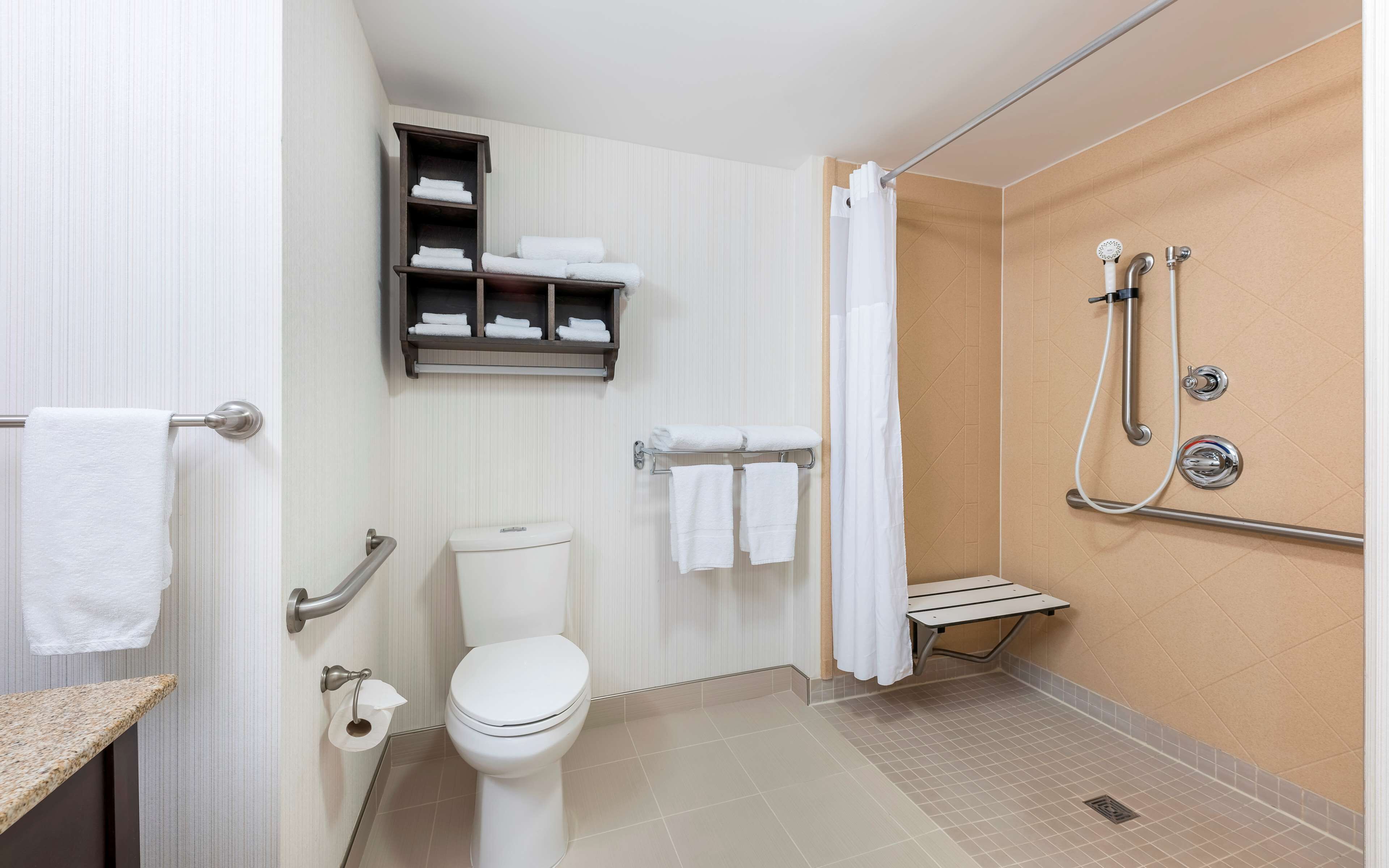 A bathroom with a toilet, a shower with a handheld showerhead, a towel rack with towels, and a shelf with folded towels.