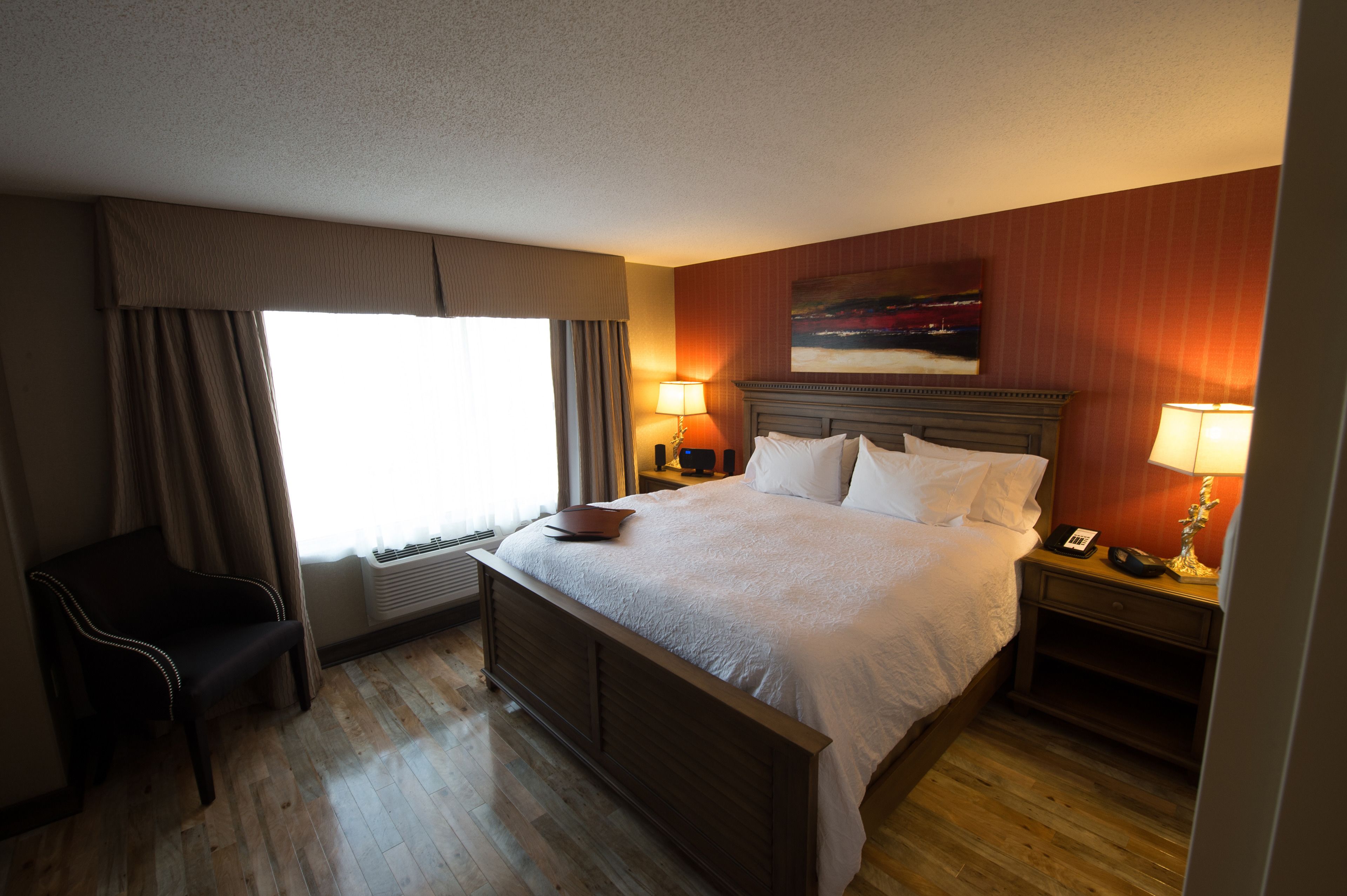 A hotel room with a large bed, two bedside tables with lamps, a chair, a window with curtains, and a painting on the wall.