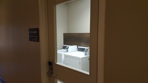 Habitación de hotel con un espejo que refleja un baño, un cartel con el horario de la lavandería para huéspedes, y una puerta con manija.