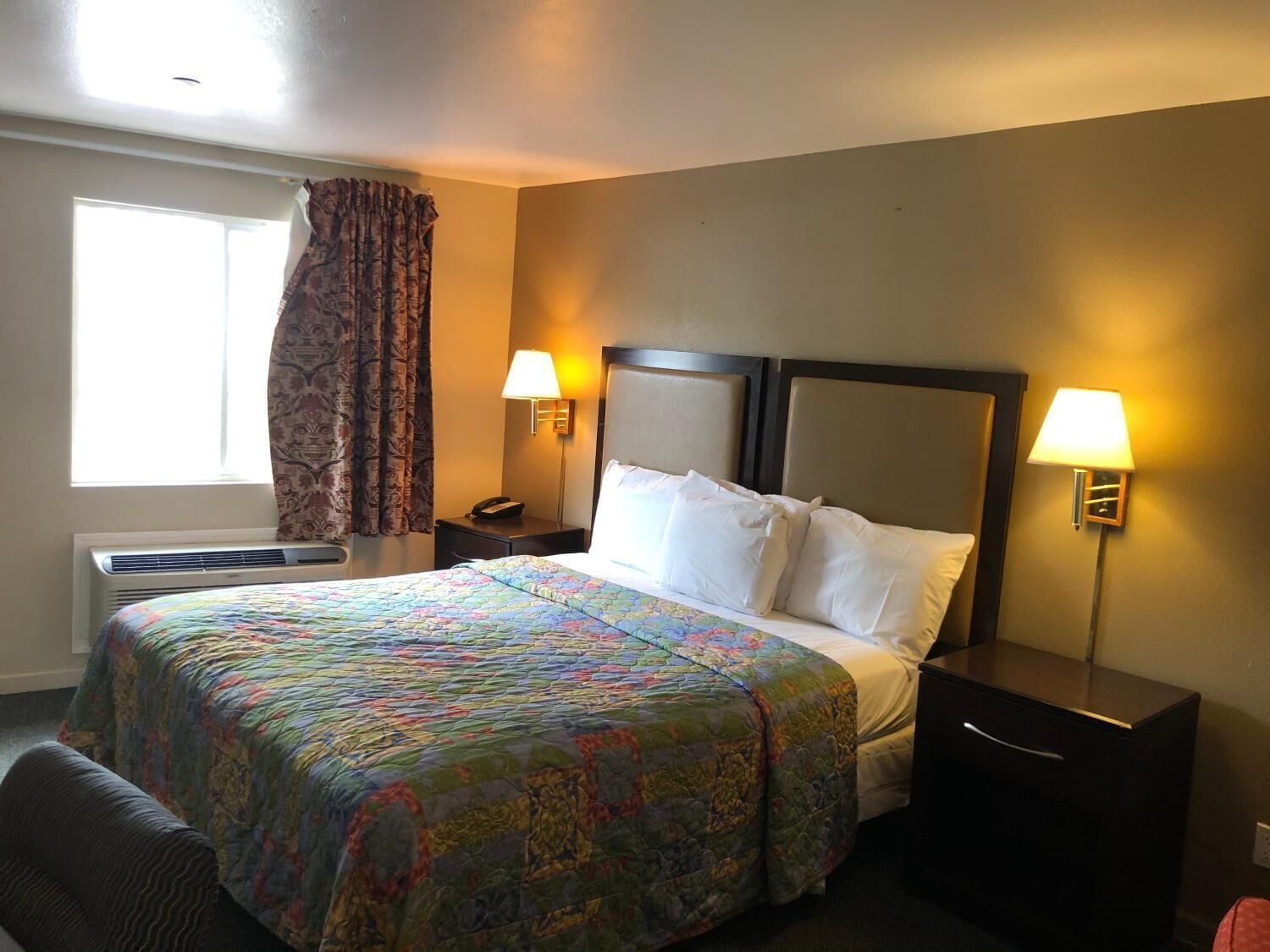 A hotel room with a bed, two bedside tables, a nightstand, a window with curtains, and a wall-mounted lamp.