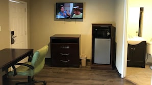 Un espacio habitable compacto con un televisor montado en la pared, una kitchenette pequeña, un comedor con mesa y silla, y una puerta que conduce a otra habitación.