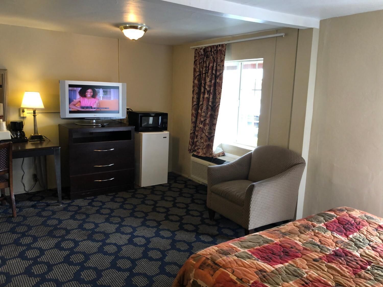 Ein Hotelzimmer mit einem Bett, einem Schreibtisch mit Fernseher, einer Mikrowelle, einem kleinen Kühlschrank, einem Sessel und einem Fenster mit Vorhängen.